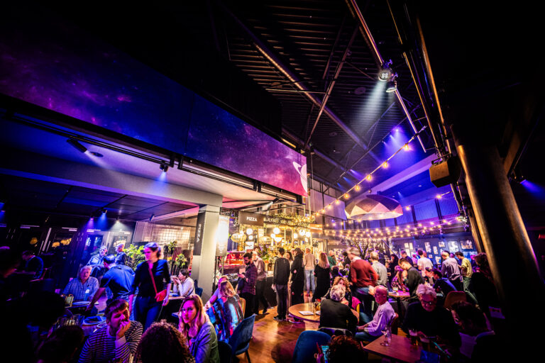 Drukke en sfeervolle bar met mensen die genieten van een avond uit, verlicht door kleurrijke lichten.