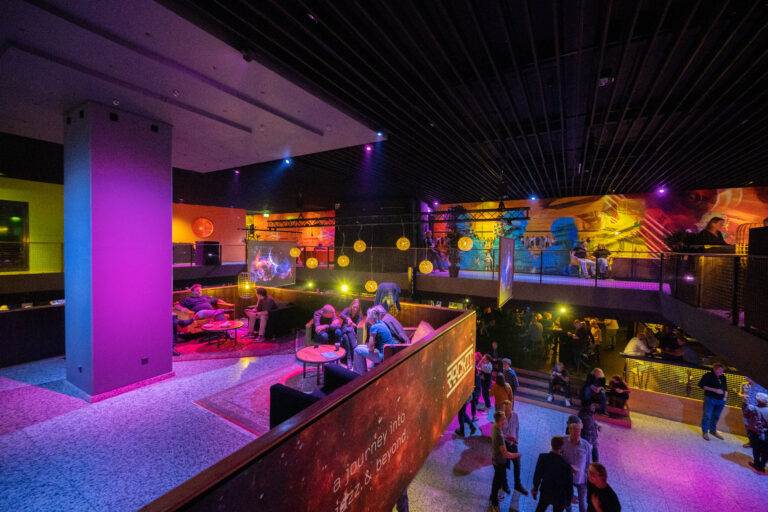 Een kleurrijke en sfeervolle lounge met verlichting in paarse en gele tinten, waar mensen bezig zijn met sociale interactie.