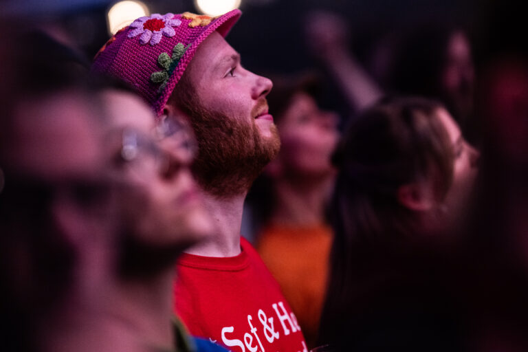 Een man met een kleurrijke hoed en een rode T-shirt kijkt vol bewondering naar een optreden in een menigte.