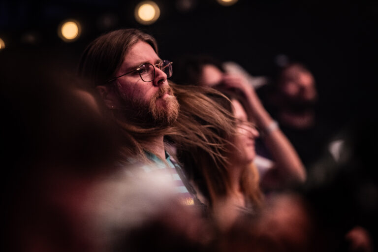 Een geconcentreerde man met lang haar en een baard, omringd door een menigte tijdens een evenement.