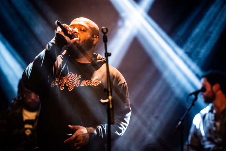 Een zanger voert op een podium zijn nummer uit, omringd door fel licht.