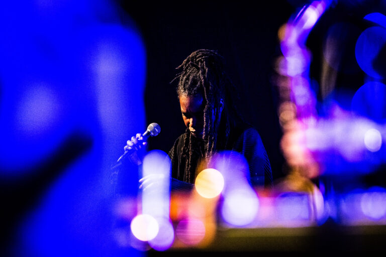 Een muzikant staat met een microfoon in een dim verlichte ruimte, omgeven door kleurrijke lichten.