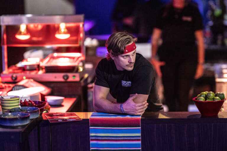 Een medewerker met een rode hoofdband leunt ontspannen op een tafel in een drukke eetomgeving.