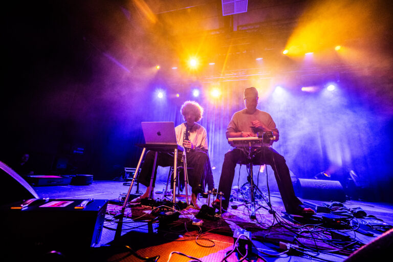 Twee muzikanten spelen live op een podium omgeven door kleurrijke lichten en rook.