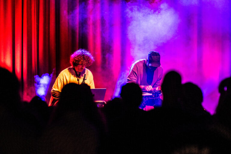 Twee muzikanten performen op een podium met kleurrijke verlichting en rookeffecten, terwijl het publiek naar hen kijkt.