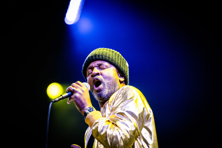 Een zanger in een gouden jas en groene muts zingt vol passie op een podium met kleurrijke lichten.