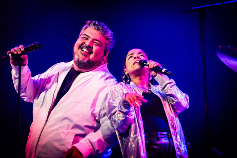 Twee performers staan samen op het podium, met een levendige achtergrondverlichting terwijl ze zingen.