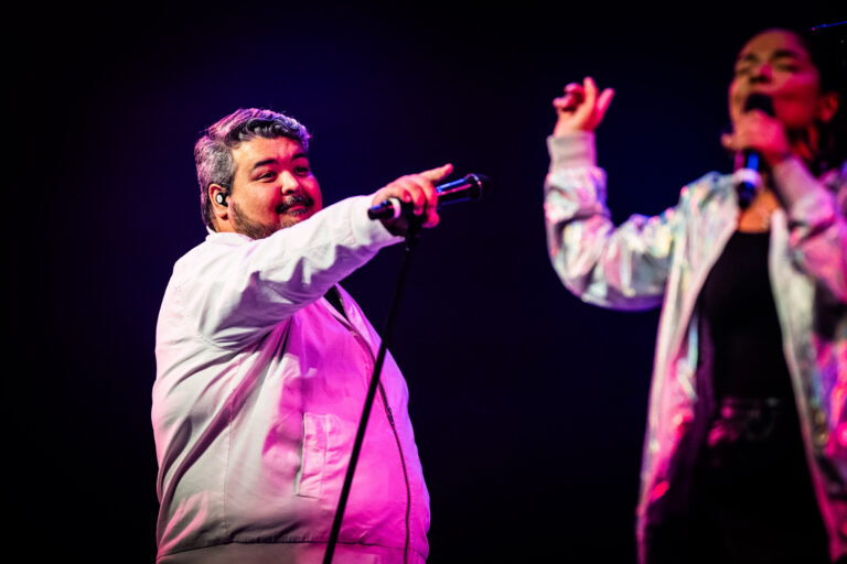 Twee performers in witte outfits staan op het podium en entertainen het publiek met een microfoon in de hand.