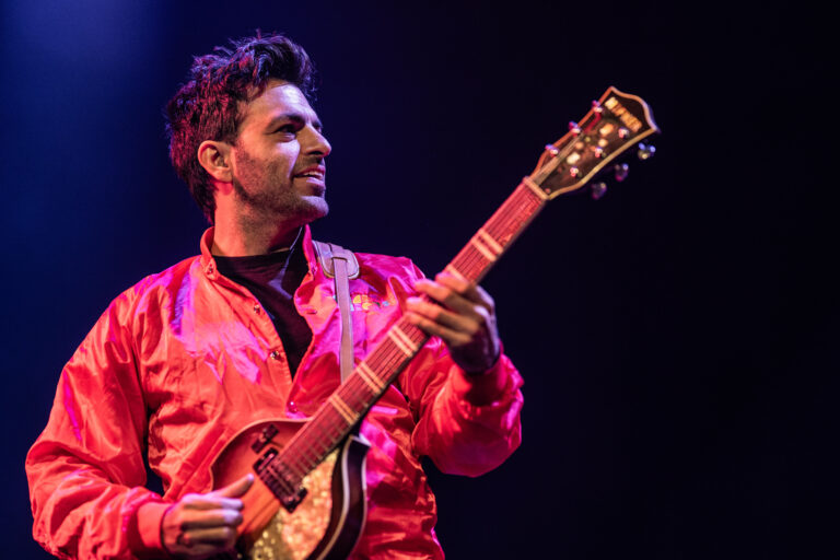 Een muzikant in een rood jack die gitaar speelt op het podium.