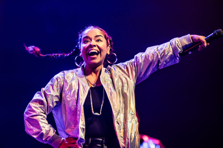 Een vrouwelijke artiest met een blijde uitdrukking steekt haar arm omhoog tijdens een optreden op een kleurrijk verlichte achtergrond.