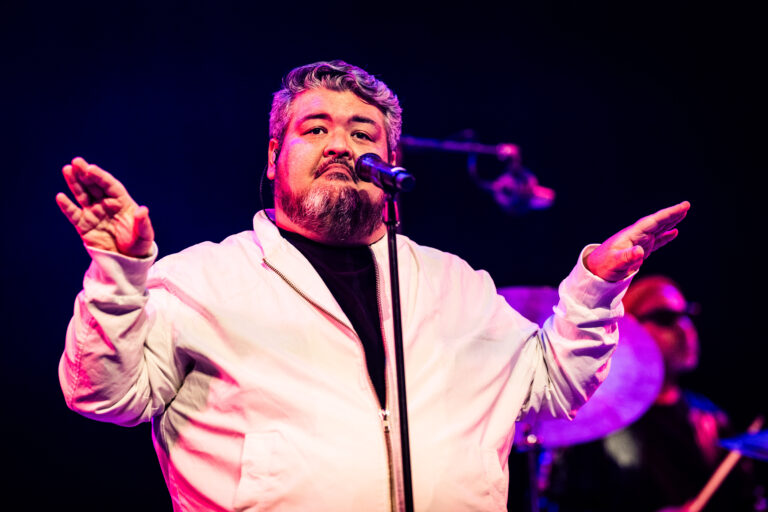 Een man met een baard zingt in een microfoon terwijl hij zijn handen gebaart op het podium.