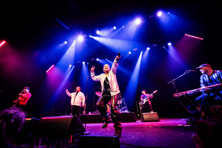 Een zanger voert op een podium met kleurrijke verlichting, omringd door muzikanten.