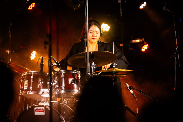 Een vrouwelijke drummer die optreedt op een podium met theatrale verlichting.