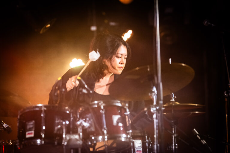 Een drummer speelt enthousiast op een drumstel met een sfeervolle lichtachtergrond.