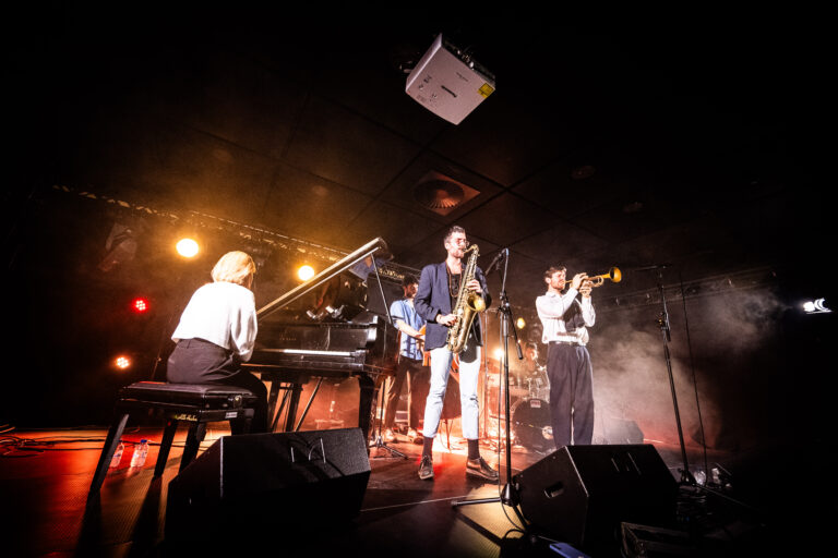 Een muzikale band speelt live op het podium met een piano, saxofoon en trompet, omringd door sfeervolle verlichting.