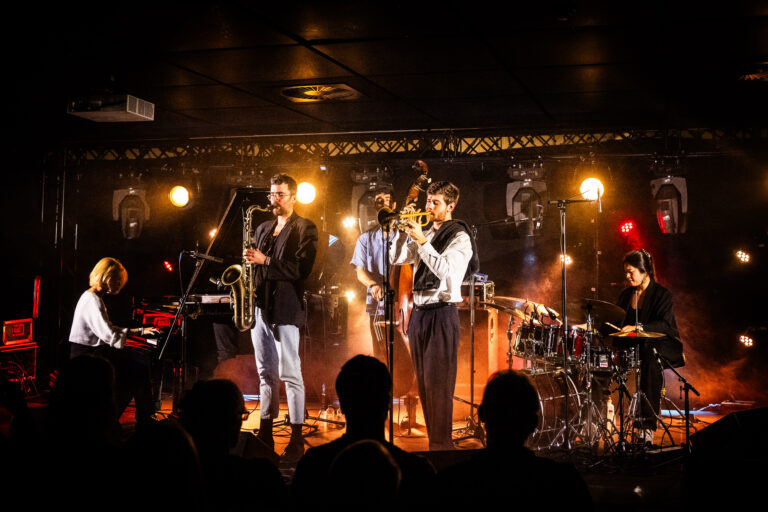 Een live band treedt op met verschillende muzikanten en instrumenten op een sfeervol verlichte podiumomgeving.