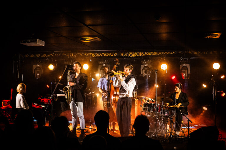 Een muzikale band voert een energiek optreden uit op een podium, omgeven door felle lichten en een enthousiast publiek.