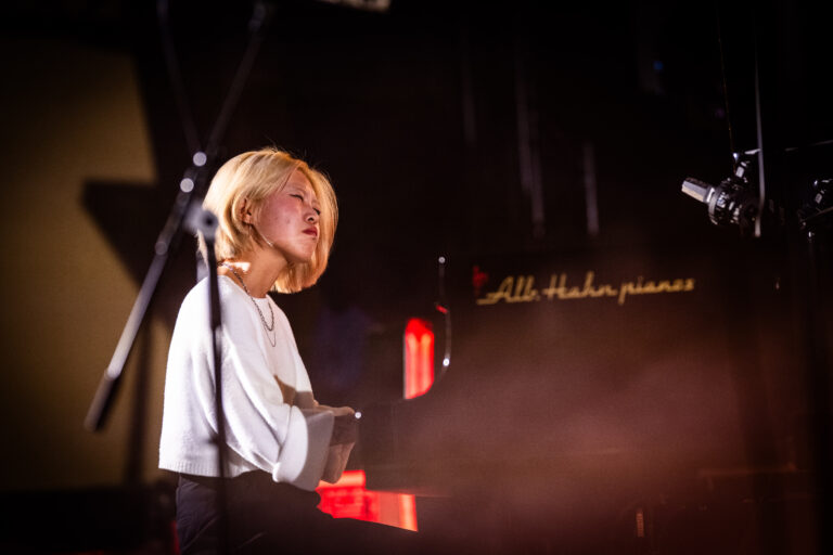 Een vrouw speelt piano op een podium, omringd door dim licht en rook.