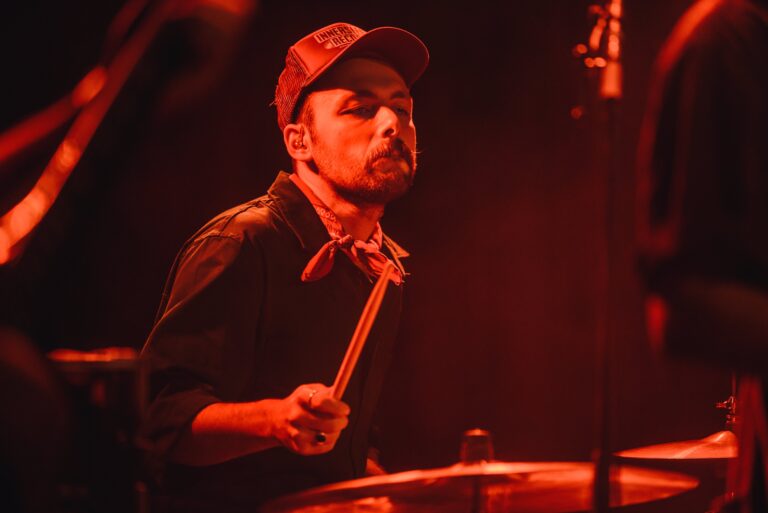 Een drummer speelt op een podium, gehuld in een warme, rode gloed.