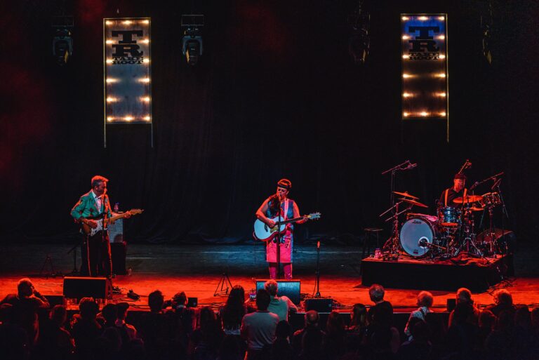 Een band speelt live op een podium met felgekleurde verlichting voor een enthousiaste menigte.