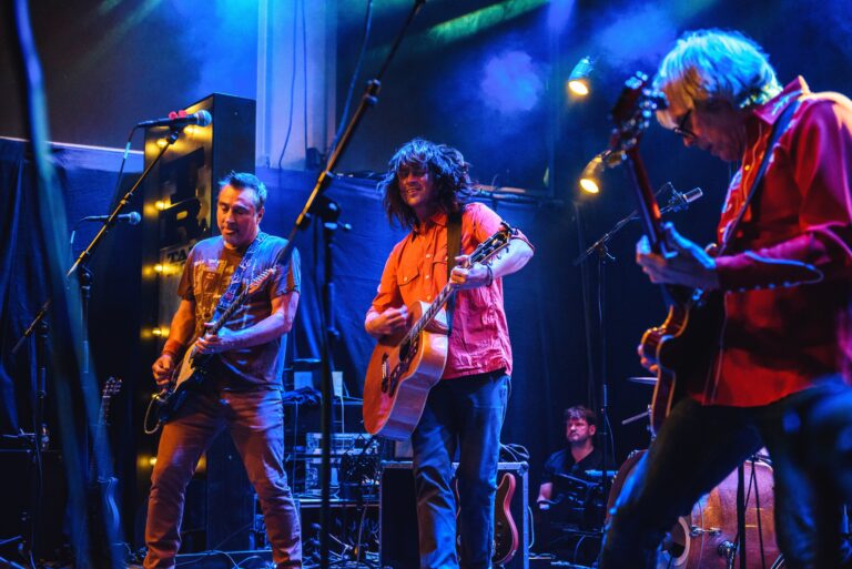 Een band speelt live op het podium met gitaren, omringd door kleurrijke lichten.