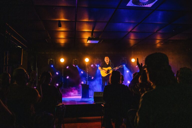Een muzikant met een gitaar staat op een podium, omringd door kleurrijke verlichting en een enthousiast publiek.