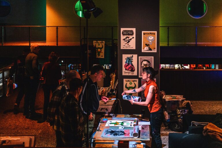 Een kunsttentoonstelling met mensen die bezoekers bedienen aan een tafel vol kunstwerken en posters aan de muur.