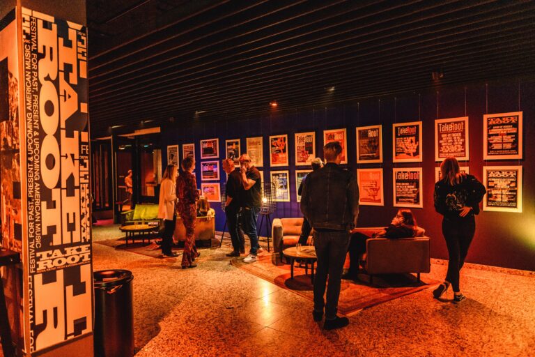 Een moderne lounge met bezoekers die voor kleurrijke posters staan, terwijl zachte verlichting een warme sfeer creëert.