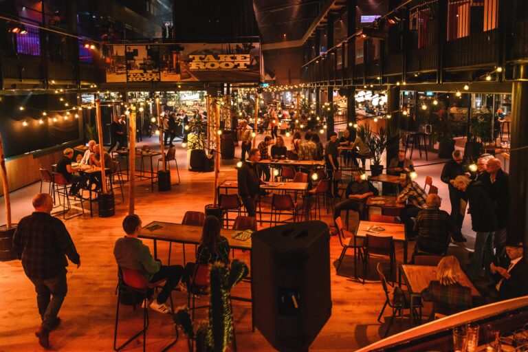 Een verlichte buitenruimte met tafels en stoelen, druk bezocht door mensen in de avond.