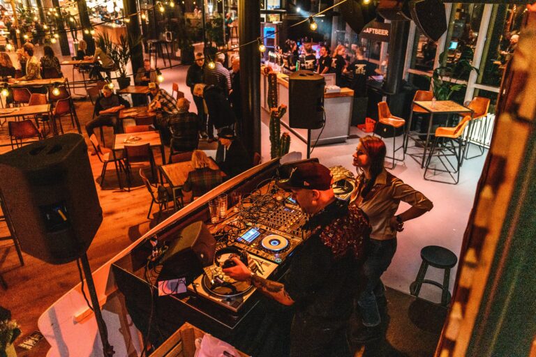 Een DJ draait op een booth in een drukke bar, omringd door gasten en sfeervolle verlichting.