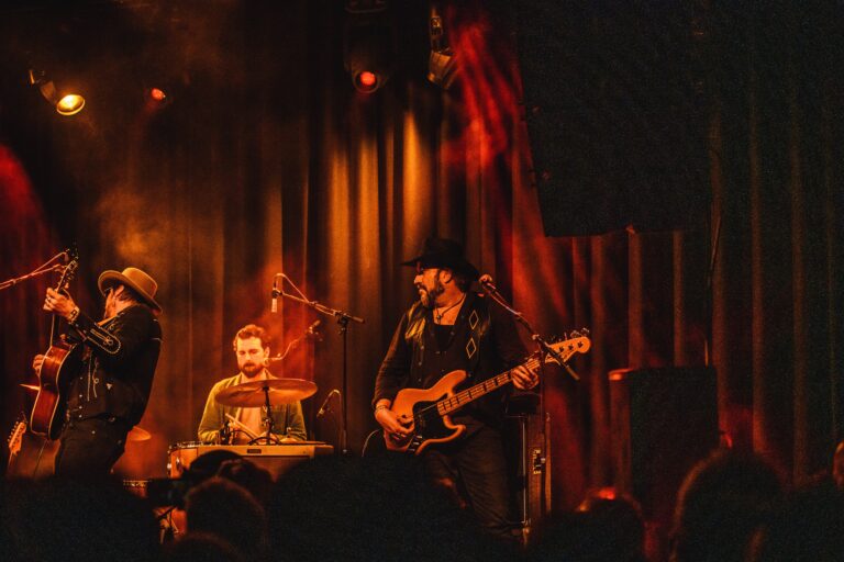 Een band speelt op het podium met een bassist en een drummer in een sfeervolle, verlichte omgeving.