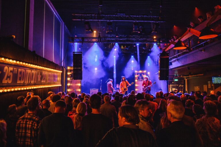 Een live band speelt op een podium met kleurrijke verlichting, terwijl een menigte naar het optreden kijkt.