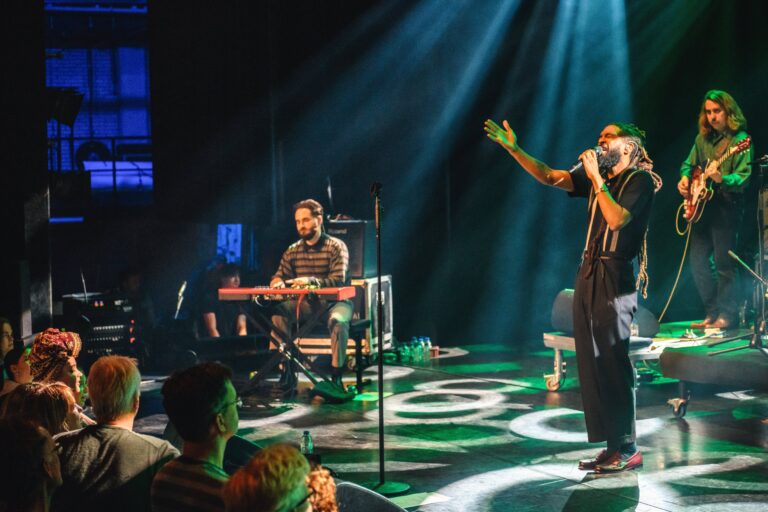 Een muzikant voert een optreden uit op een podium, terwijl een andere muzikant op de achtergrond speelt op een keyboard.