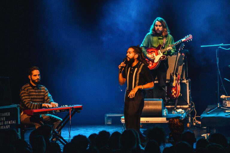 Een muzikant staat centraal op het podium terwijl een andere muzikant op een keyboard speelt en een derde op de achtergrond gitaar speelt.