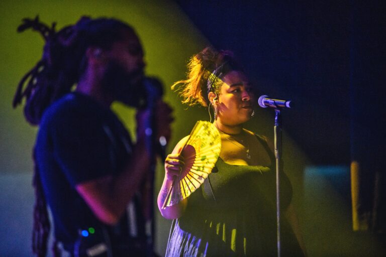 Twee muzikanten staan op het podium, een zangeres met een microfoon en een man die achtergrondzang of instrument speelt, omlijst door kleurrijke lichten.