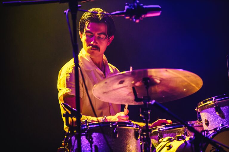 Een drummer speelt enthousiast op een set drums onder kleurrijke belichting.