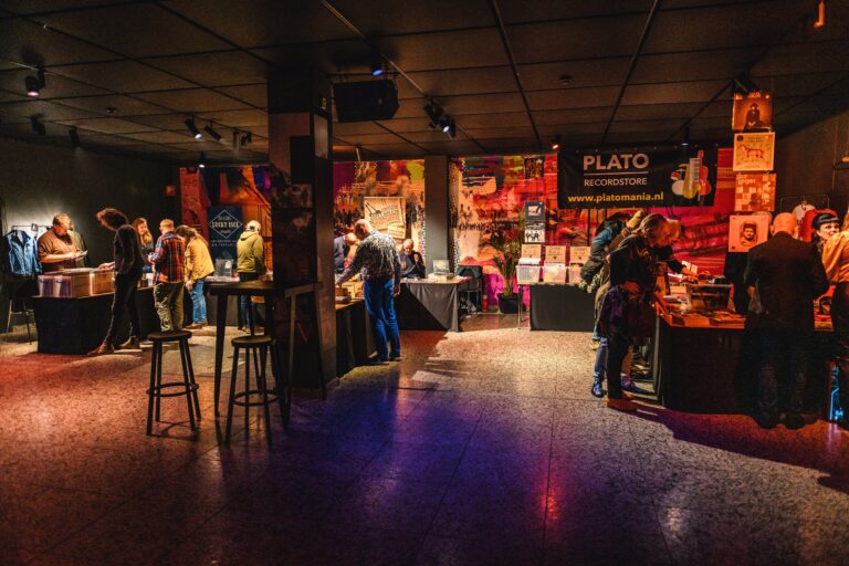 Een drukke binnenruimte met mensen die rond circuleren en verschillende standen en tafels bekijken.