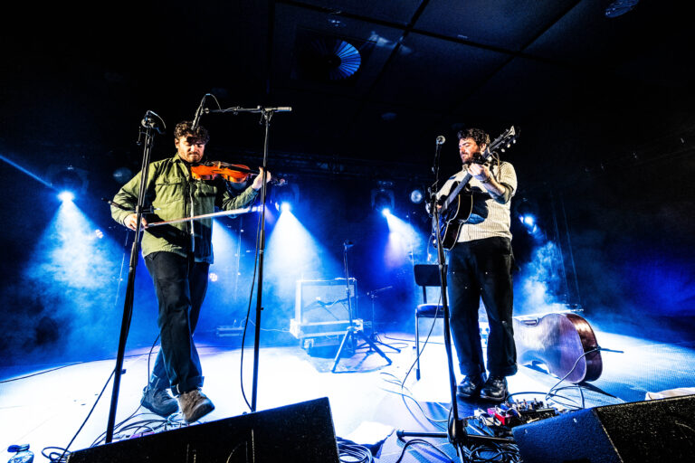 Twee muzikanten optreden op een podium, met een viool en een gitaar, omgeven door blauwe lichten en rook.
