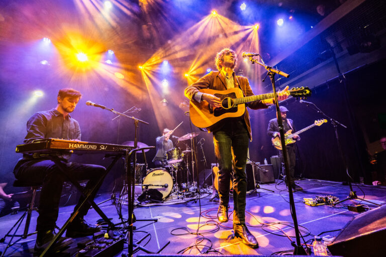 Een muzikant met een gitaar staat op het podium, omringd door bandleden en kleurrijke lichten.