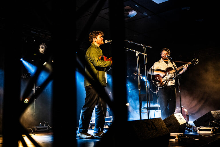 Een liveconcert met twee muzikanten op het podium, omringd door blauwe belichting.