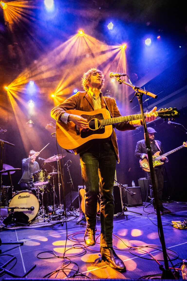 Een muzikant speelt op een gitaar terwijl hij optreedt op een podium met kleurrijke lichten.