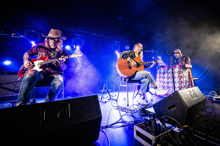 Drie muzikanten spelen op het podium met een gitaar, omringd door kleurrijke verlichting en een sfeervolle rook.