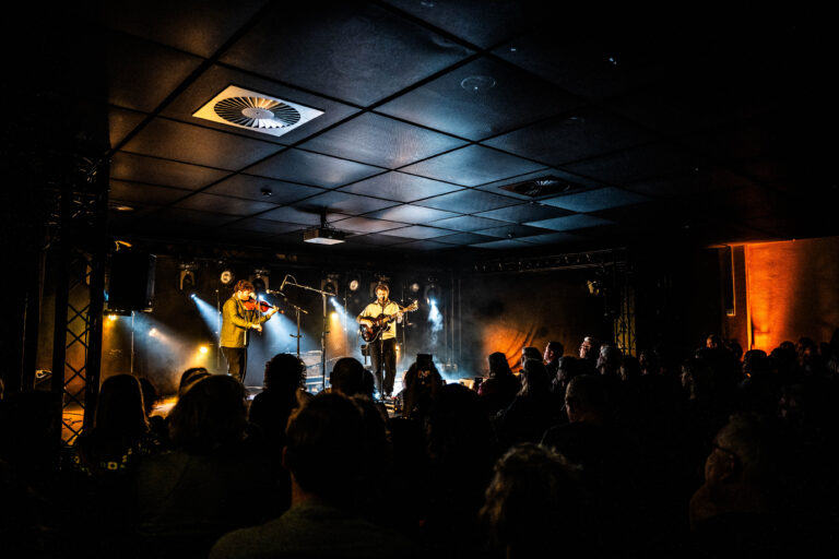 Een band speelt live op het podium voor een publiek in een sfeervolle, donkere ruimte met theatrale belichting.
