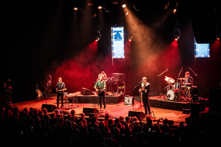 Een band speelt live op het podium met kleurrijke verlichting en een enthousiast publiek.