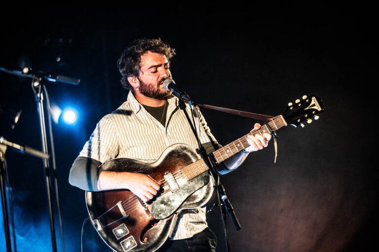 Muzikant die in een dimverlichte omgeving speelt op een gitaar, terwijl hij zingt in een microfoon.