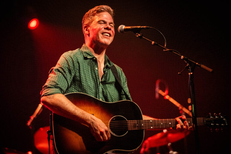 Een muzikant zingt en speelt gitaar op het podium met een aanwezige lichtshow.