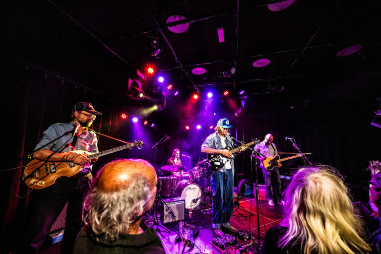 Een band speelt op een podium met kleurrijke verlichting, terwijl het publiek naar hen kijkt.