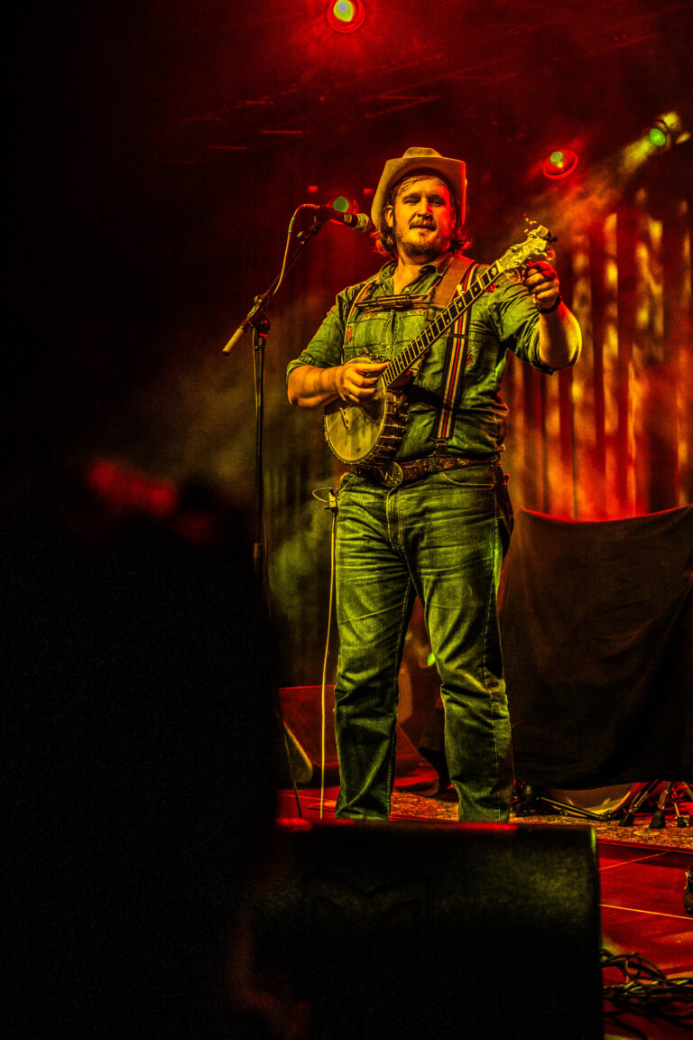 Een muzikant speelt op een banjo onder kleurrijke verlichting op een podium.