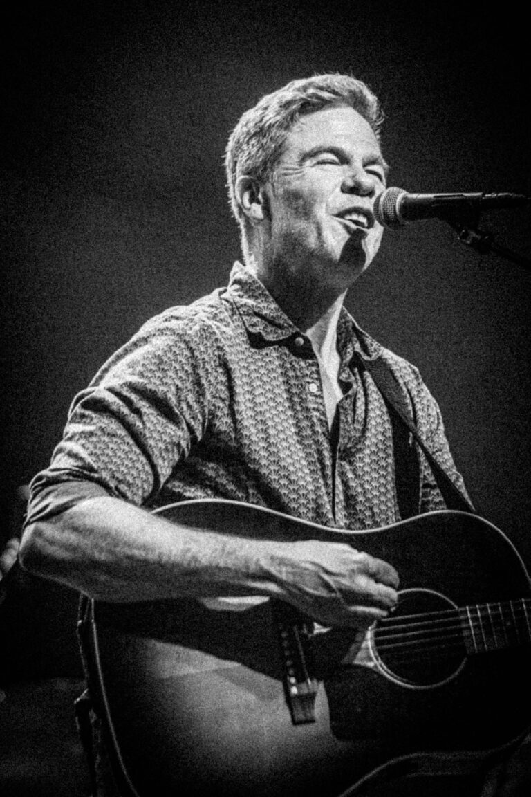 Een man met een gitaar zingt in een microfoon, verlicht door een spotlicht tegen een donkere achtergrond.