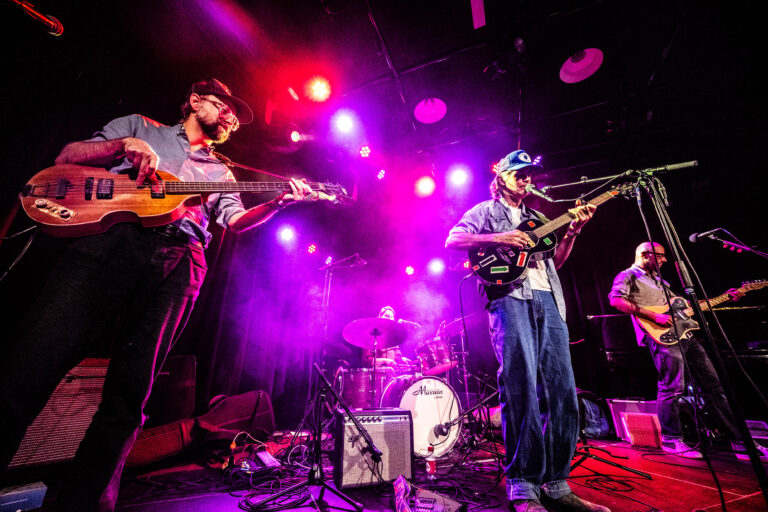 Muzikanten treden op op een live podium, omringd door kleurrijke lichten en rook.